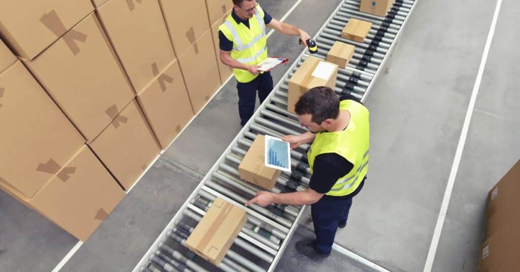 Two men in bright yellow vests and in a warehouse setting scan items on tablets as boxes move down a conveyor belt, Are part time workers covered by workers comp.jpg