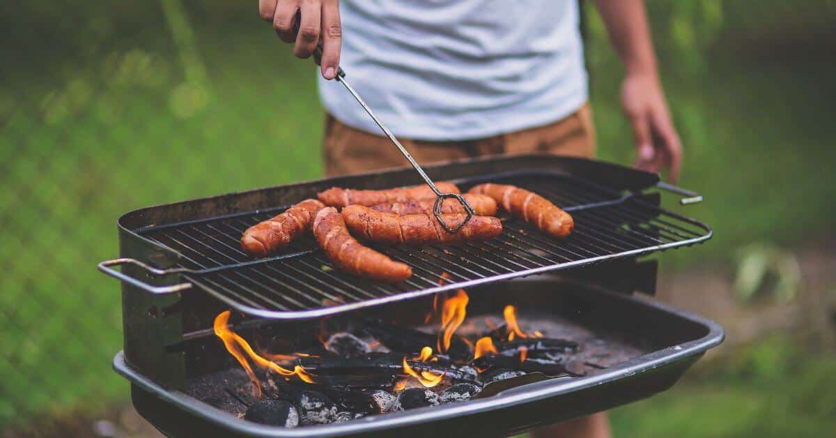 A man holds tongs over an outdoor flame grill filled with hot dogs, summer injuries, personal injury attorneys